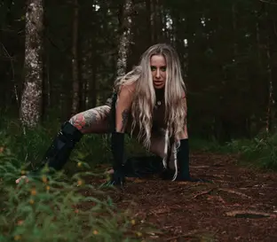 Melanie crouches on a forest path, wearing black gloves and boots, staring at camera.