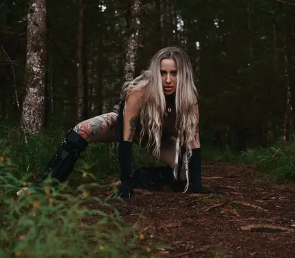 Melanie crouches on a forest path, wearing black gloves and boots, staring at camera.